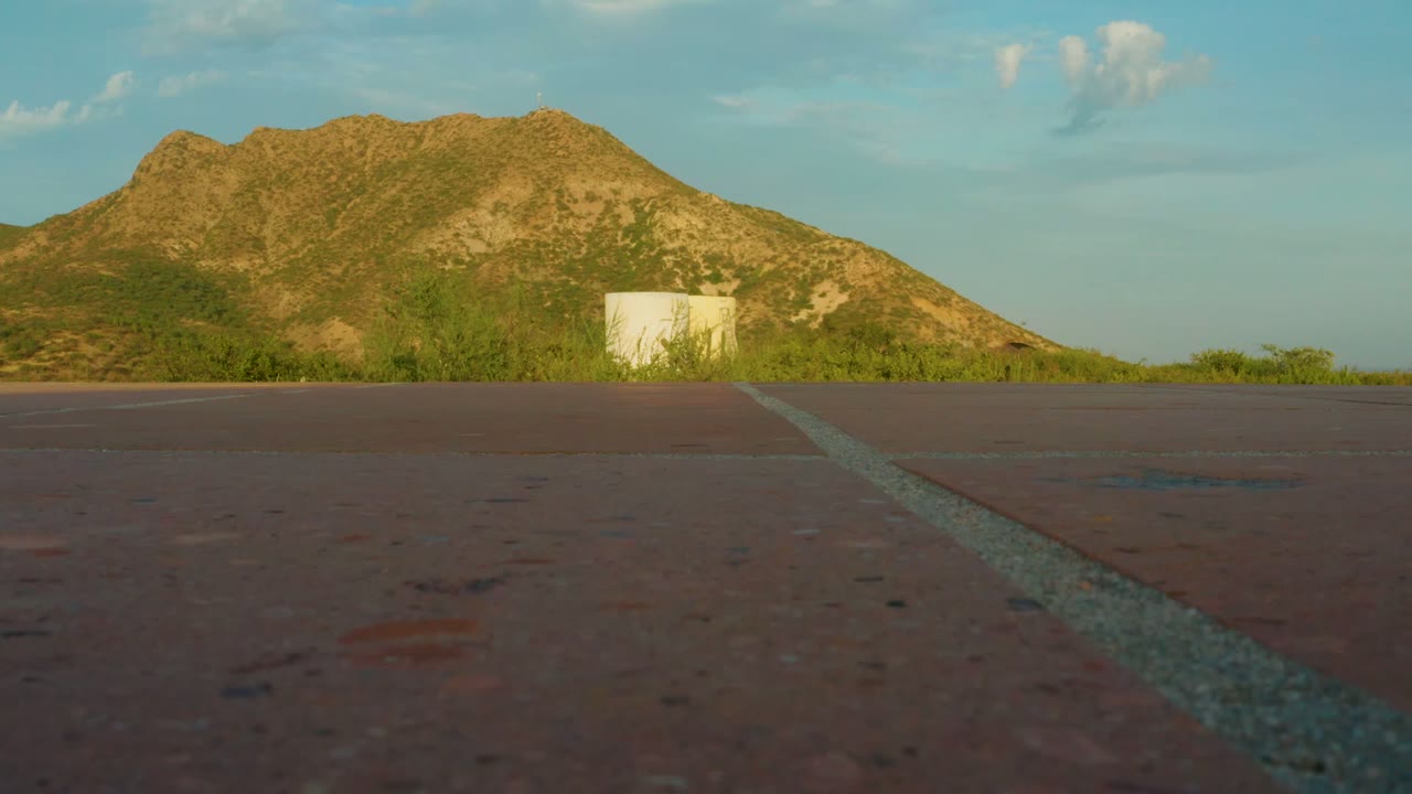 At 0:02, the road remains low in the foreground while the small white structure sits right of center below the sunlit mountain, with sparse clouds overhead and no visible people.
