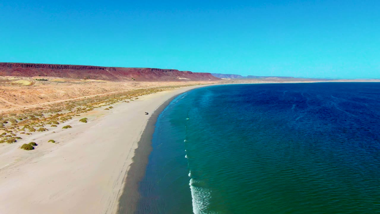 Aerial frame over San Juanico, Baja California Sur showing a small dark vehicle on pale sand, turquoise water along the right edge, desert scrub and red-brown bluffs inland, and no visible people.