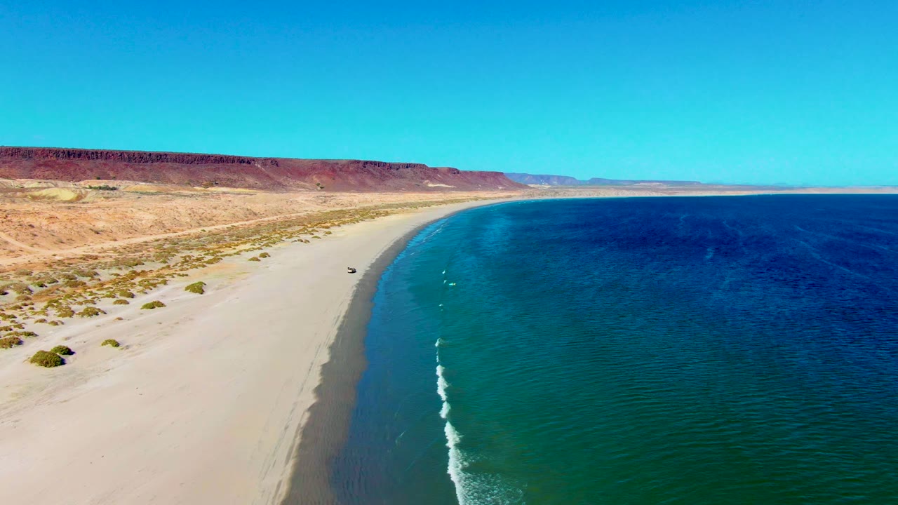 Drone view of a beach vehicle near the waterline with desert dunes and blue sea.