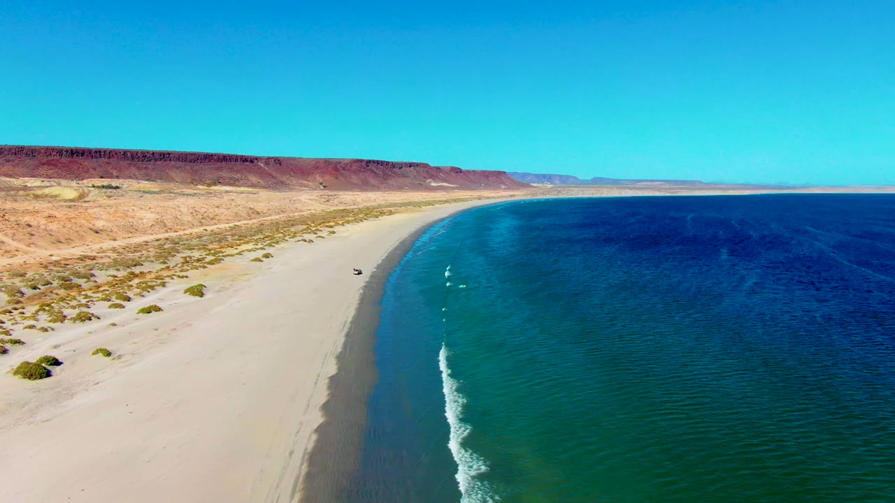 San Juanico, Baja California Sur aerial frame showing the vehicle near the left side of the shoreline curve, two small white wave marks in the blue-green water, desert flats, long bluffs, and no visible people.