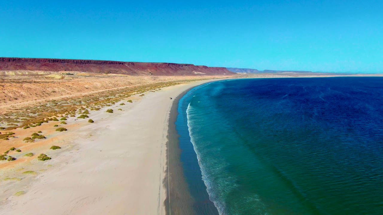 Aerial view over San Juanico, Baja California Sur, with pale sand, sparse desert scrub, a curving shoreline, deep blue water, and no visible people.