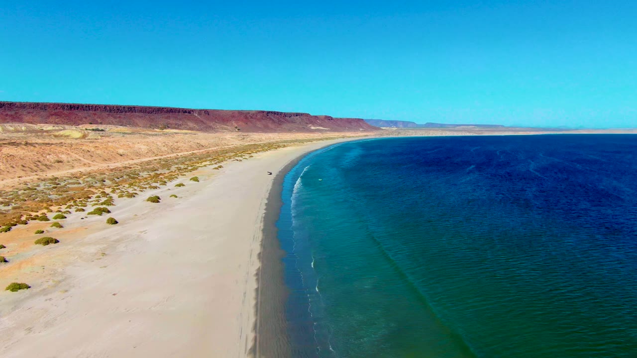 The San Juanico coastline curves into the distance as a vehicle sits near the sandy shore, with darker blue water offshore and no visible people.