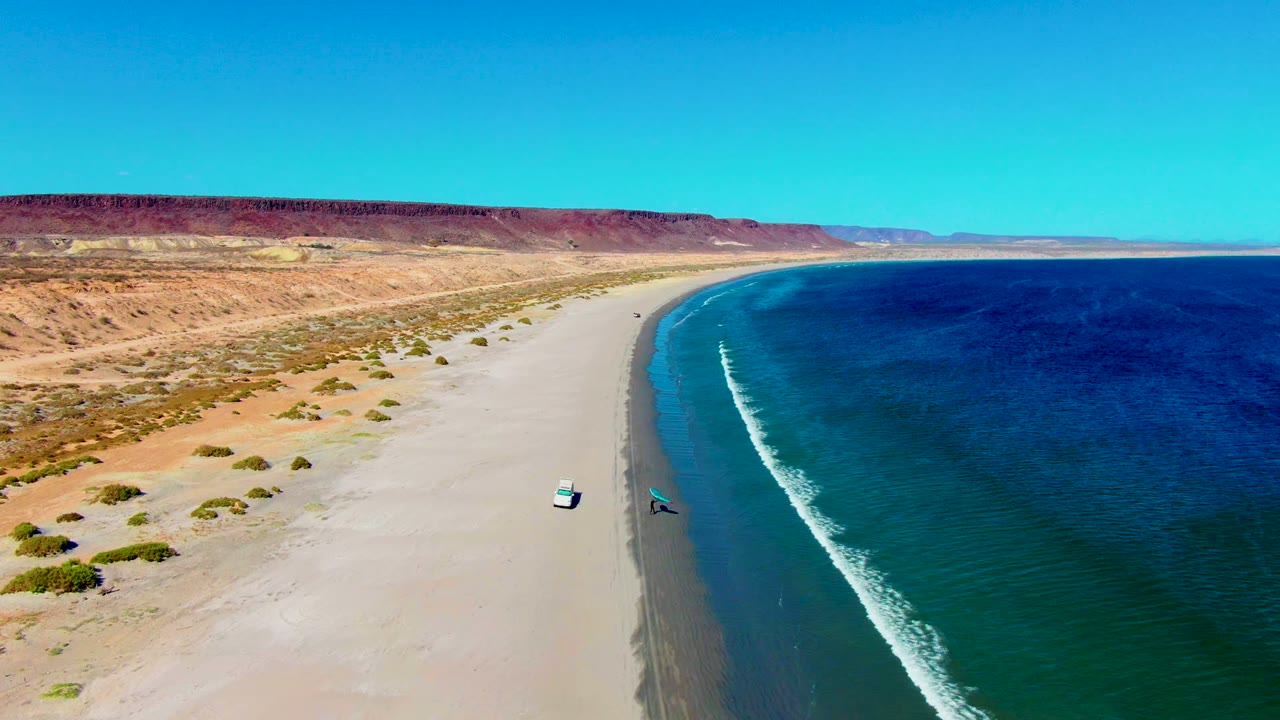 White vehicle and blue kite near the surf on a broad desert beach.