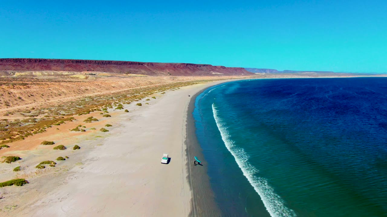 San Juanico aerial frame with the vehicle and blue kite separated on the beach, deeper blue ocean filling the right side, pale sand and low desert vegetation on the left, and no visible people.