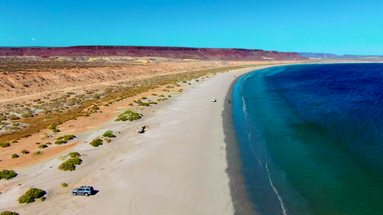 Aerial frame over San Juanico, Baja California Sur showing a pale sandy beach beside deep blue ocean, red cliffs inland, sparse shrubs, a small vehicle near the lower left, and no visible people.