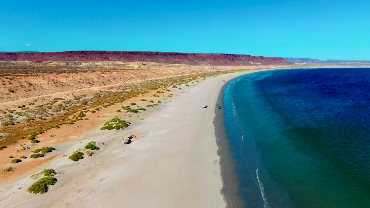 Aerial frame over San Juanico with the white vehicle farther along the sand, clear blue water curving at right, low desert dunes and shrubs at left, and no visible people.