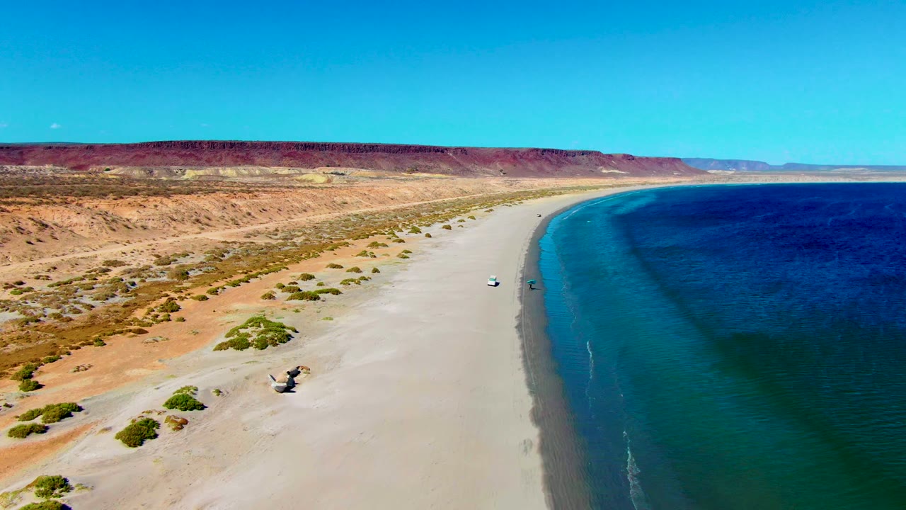 San Juanico aerial still with the beach sweeping into the distance, the white car near mid-beach, a small darker object lower on the sand, blue ocean at right, and no visible people.