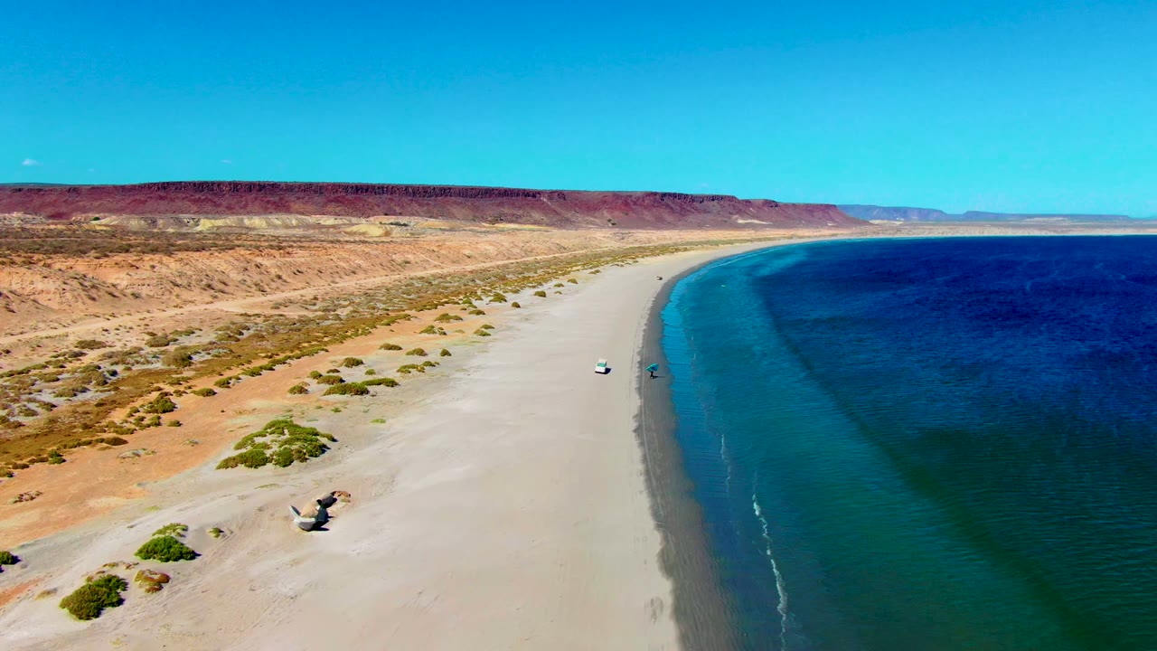 Aerial beach scene with dunes, shrubs, red cliffs, blue water, and small vehicles on sand.