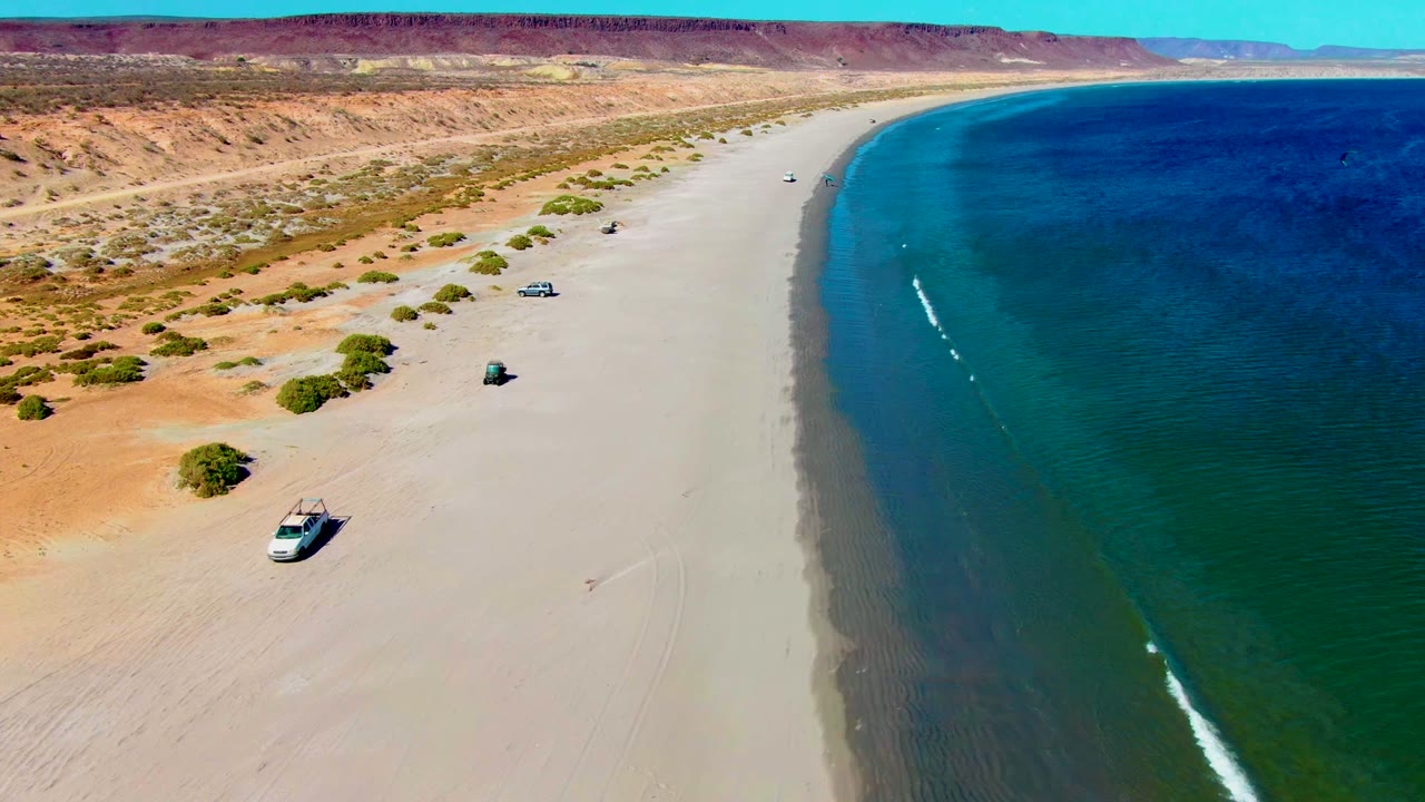 Aerial view over San Juanico, Baja California Sur, with vehicles spaced along a wide sandy beach, blue ocean at right, desert scrub and bluffs at left, and no visible people.