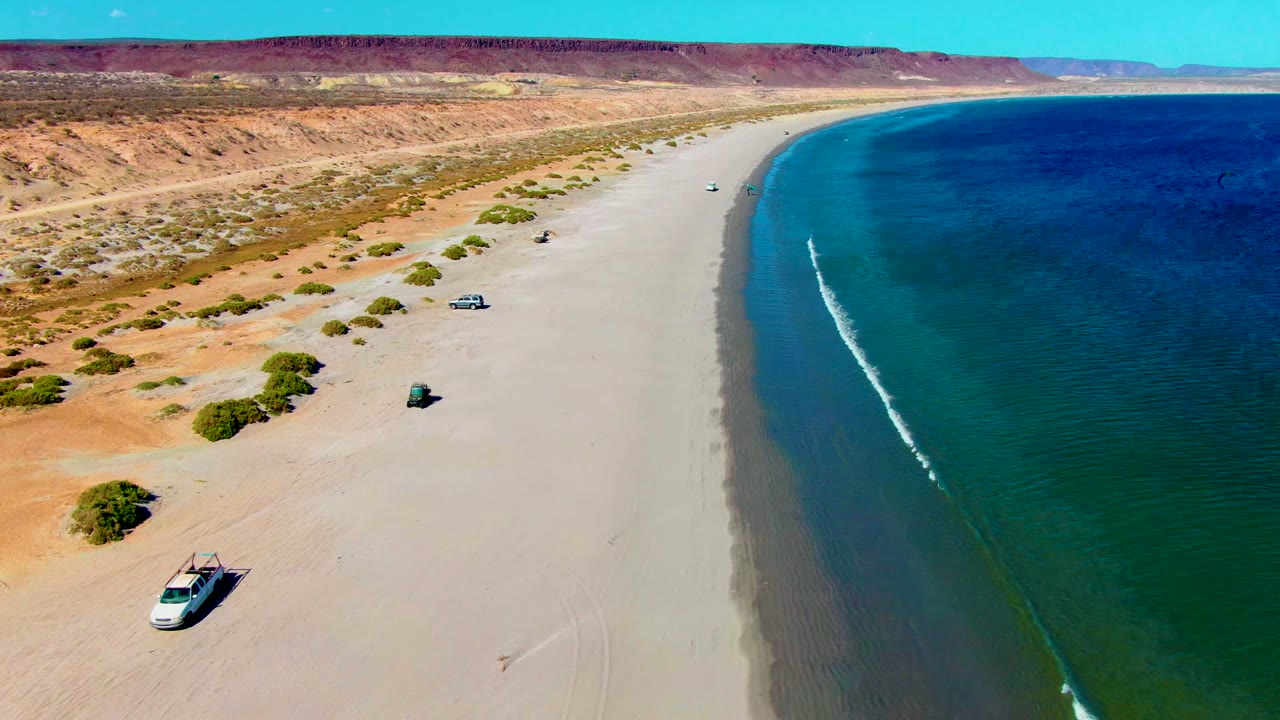 Aerial beach view with vehicles, desert scrub, bluffs, and a narrow wave line.