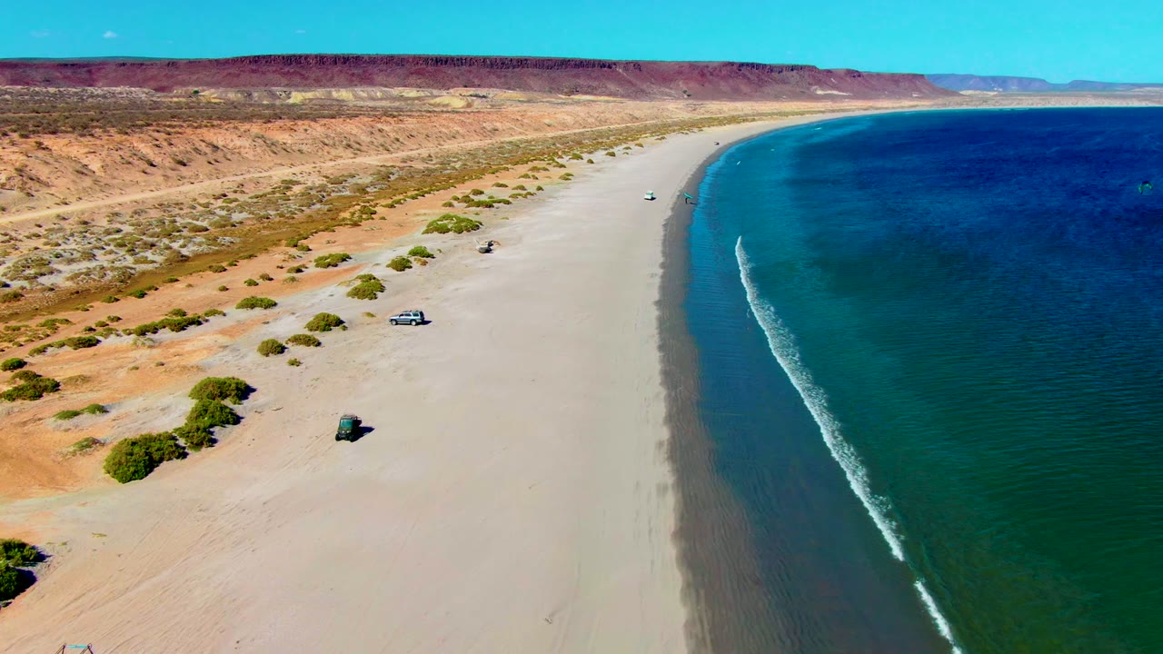 San Juanico aerial frame with a broad empty beach curving toward the distance, small vehicles on the sand, deep blue water to the right, desert bluffs inland, and no visible people.
