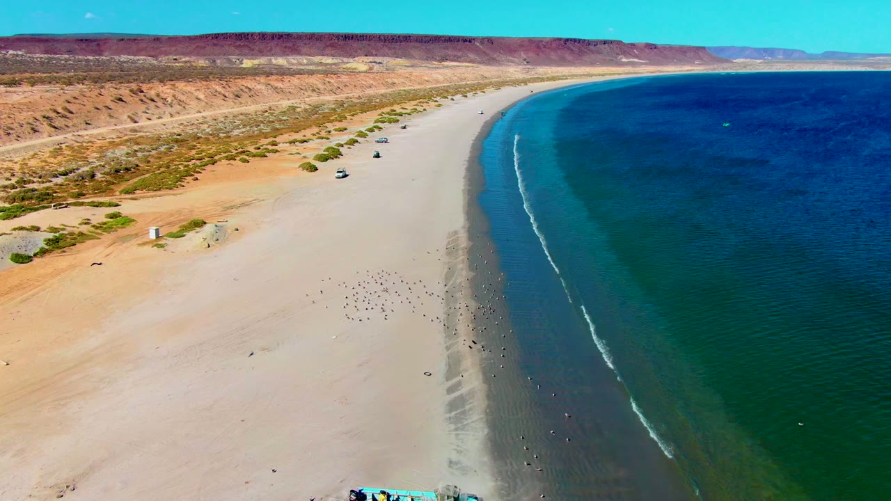 High aerial view over San Juanico, Baja California Sur, with pale sand curving along blue-green water, reddish desert mesas inland, scattered vehicles on the beach, and no visible people.