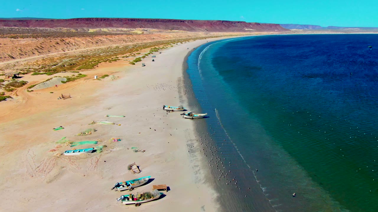 Aerial view of San Juanico, Baja California Sur, Mexico, with small boats on pale sand, clear blue-green water, desert dunes, and tiny people visible along the shoreline.