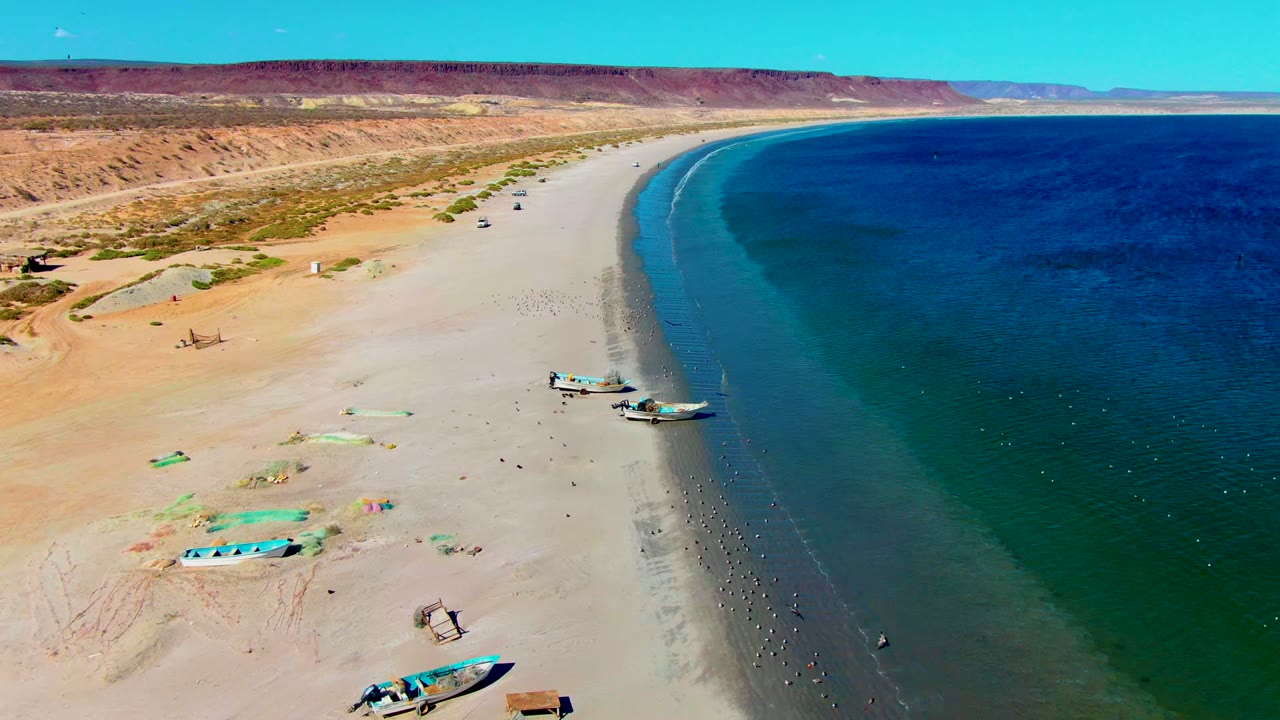 Aerial frame over San Juanico showing boats near the waterline, beach gear on the sand, scattered people, desert dunes, and the curving blue-green shoreline.