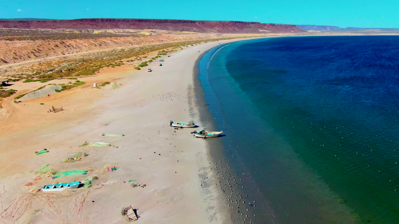 Drone view at San Juanico with two boats closest to the surf, open sand, small people, desert dunes, low cliffs, and dark blue water farther offshore.