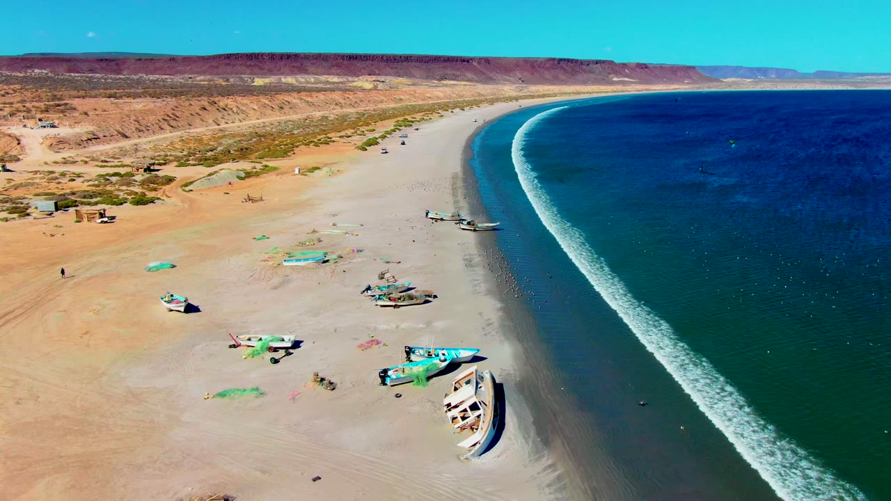 Aerial frame over San Juanico, Baja California Sur, showing several small boats on pale sand, a curving wet shoreline, blue-green ocean, desert bluffs, clear sky, and no visible people.