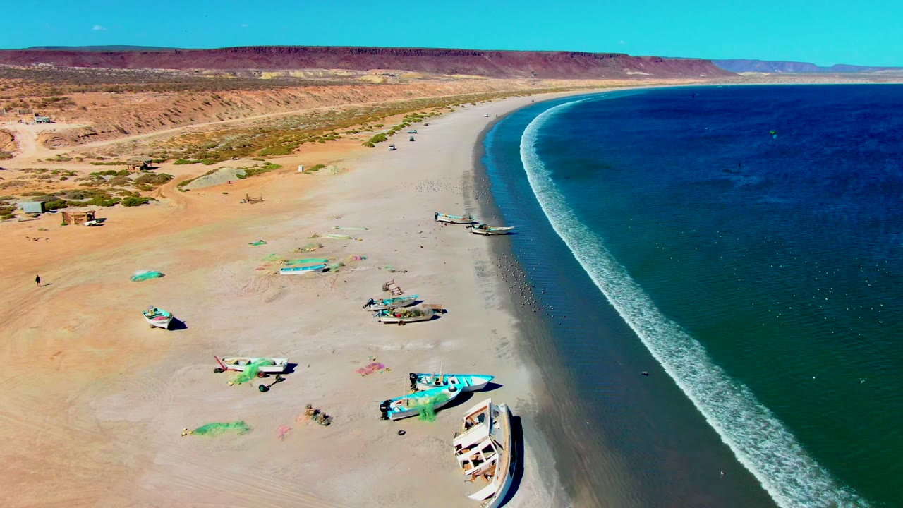 Drone view of beach boats, desert sand, and a white wave line.
