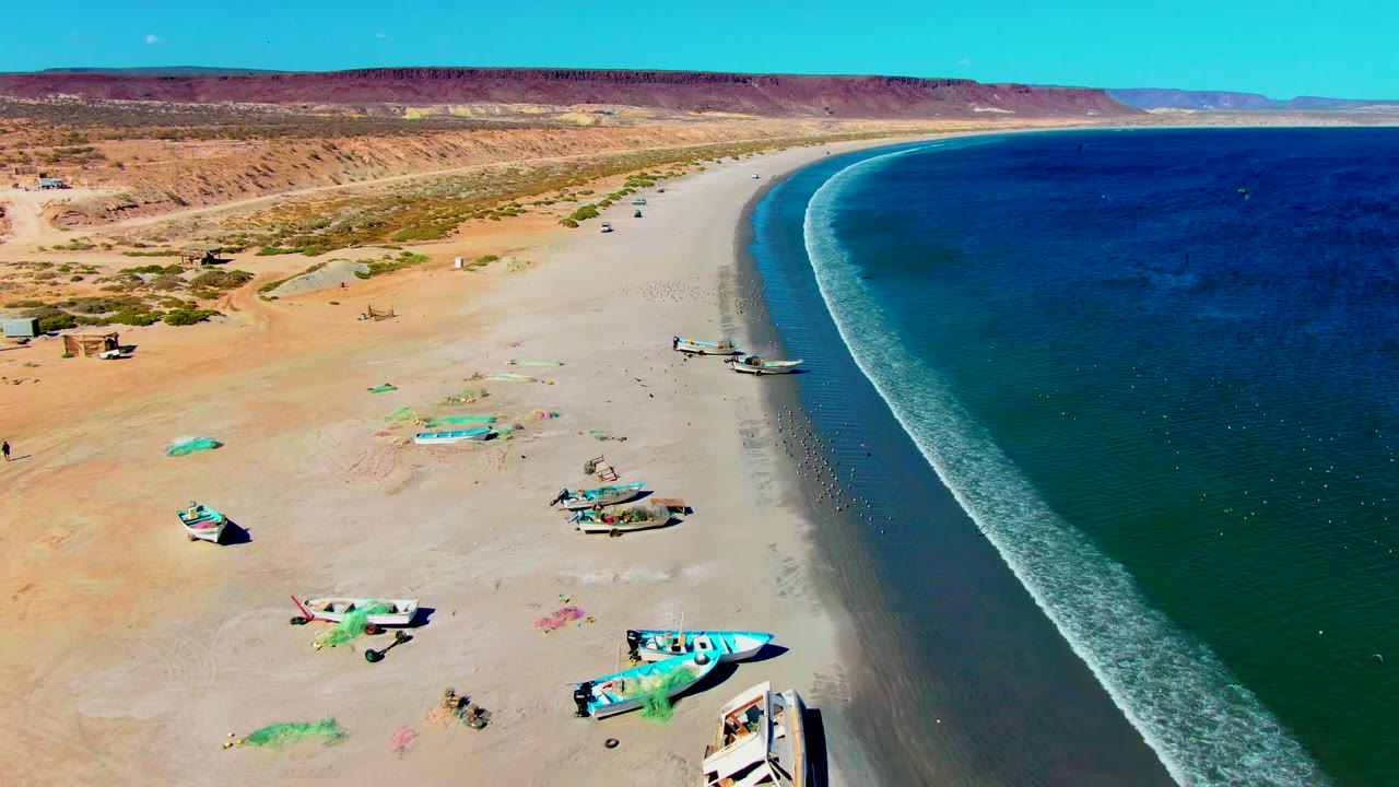 San Juanico aerial frame showing more open sand around the boats, a darker band of wet beach, deep blue water to the right, low desert hills in the distance, and no visible people.