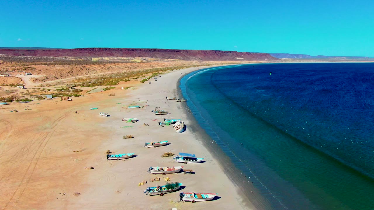 Aerial view over San Juanico, Baja California Sur, with small boats beached on pale sand, dark blue ocean along the right side, desert bluffs in the distance, and no visible people.