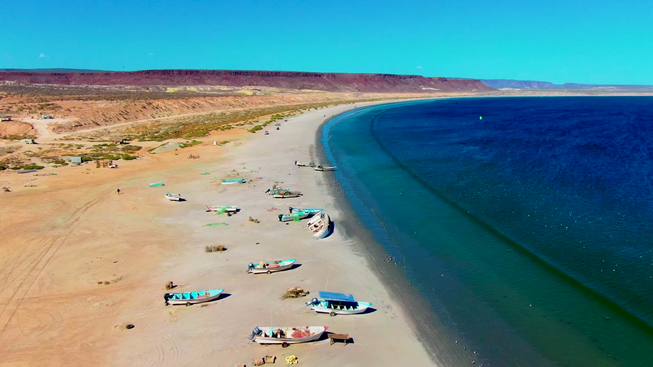 Aerial view of the San Juanico beach with scattered small boats on the sand, a narrow wet shoreline band, calm blue water to the right, desert bluffs beyond, and no visible people.