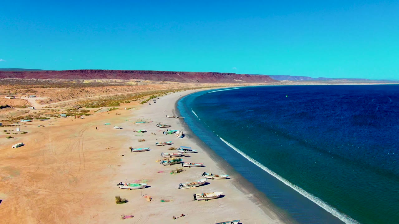 Aerial view in San Juanico, Baja California Sur, with fishing boats hauled onto pale sand, blue ocean at right, desert ridge in the distance, and tiny people visible near the boats.