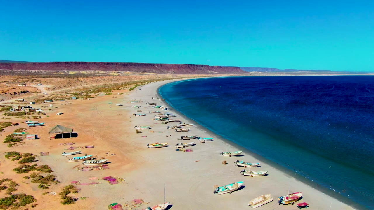 Aerial frame over San Juanico, Baja California Sur showing fishing boats spaced along pale sand, deep blue water on the right, desert hills beyond, and no visible people.