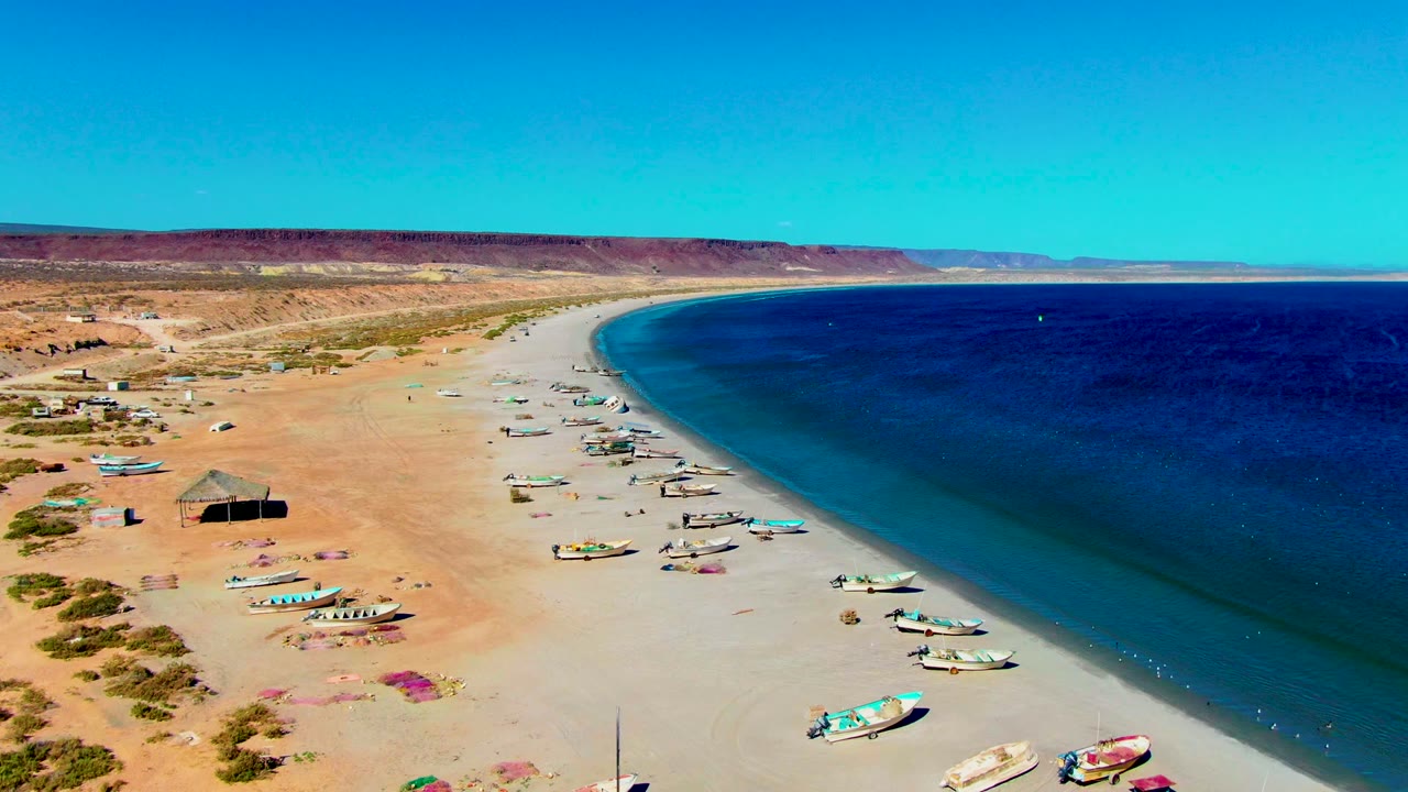 Fishing boats sit along a curved sandy shore with desert hills behind.