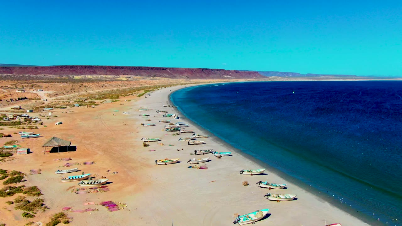 High aerial frame over San Juanico, Baja California Sur showing fishing boats clustered near the waterline, a wide empty sand area, dark blue ocean, desert background, and no visible people.