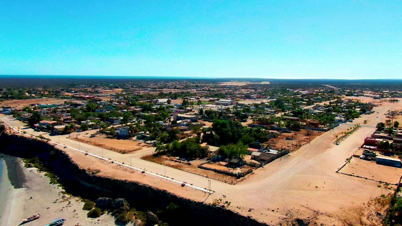 Aerial view over San Juanico, Baja California Sur, with a sandy beach at left, a dirt road along the bluff, low homes and trees beyond, and no visible people.