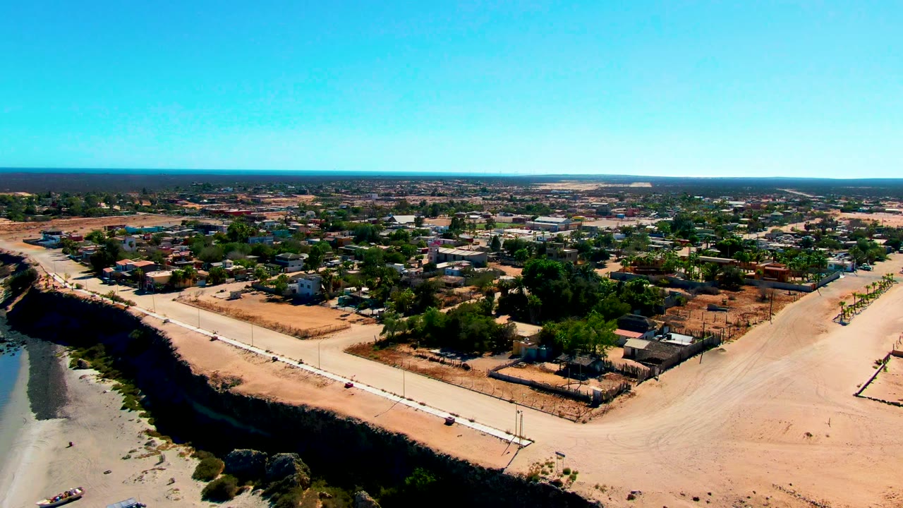 Wide aerial frame over San Juanico with dry lots and trees behind a light dirt road, the beach and shallow waterline along the left edge, and no visible people.