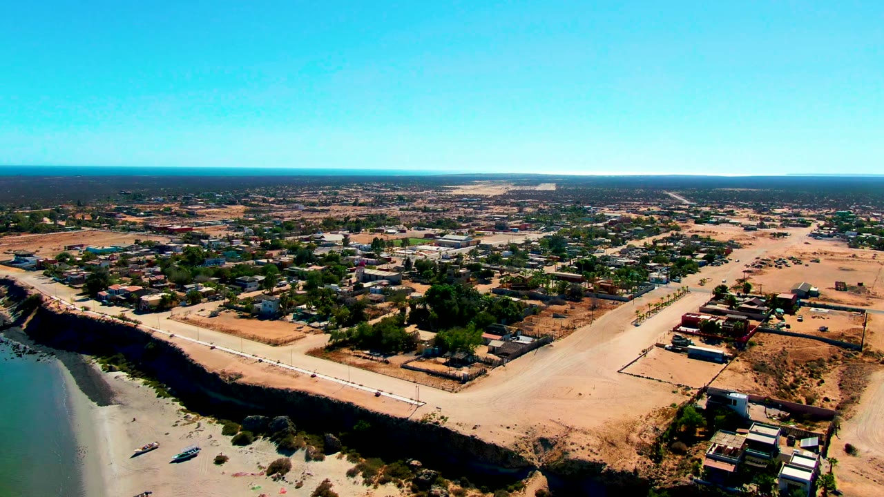 Aerial frame over San Juanico, Baja California Sur showing calm water, a sandy beach with small boats, a dirt road, scattered buildings, dry lots, and no visible people.