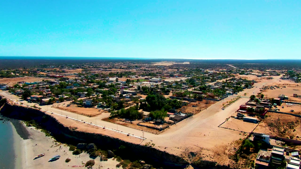 Drone frame over San Juanico showing small boats pulled near the sandy shore, a dark bluff edge, low buildings, open dry ground, a broad road, and no visible people.