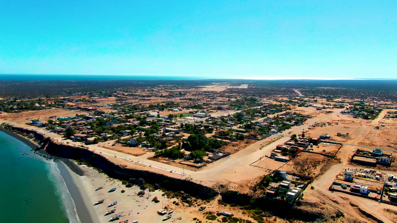 Aerial frame over San Juanico, Baja California Sur, showing green-blue ocean, a sandy beach with small boats, a road above the bluff, dry lots, low buildings, and no visible people.