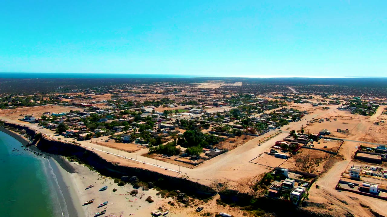 Wide aerial frame in San Juanico showing the beach and boats at lower left, the road across the center, low buildings and sandy lots to the right, and no visible people.