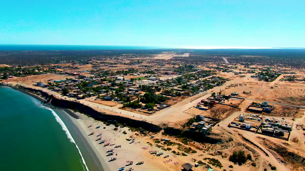 Aerial frame over San Juanico, Baja California Sur, Mexico, with turquoise ocean at left, small boats on the sandy beach, dry roads and buildings inland, and no visible people.