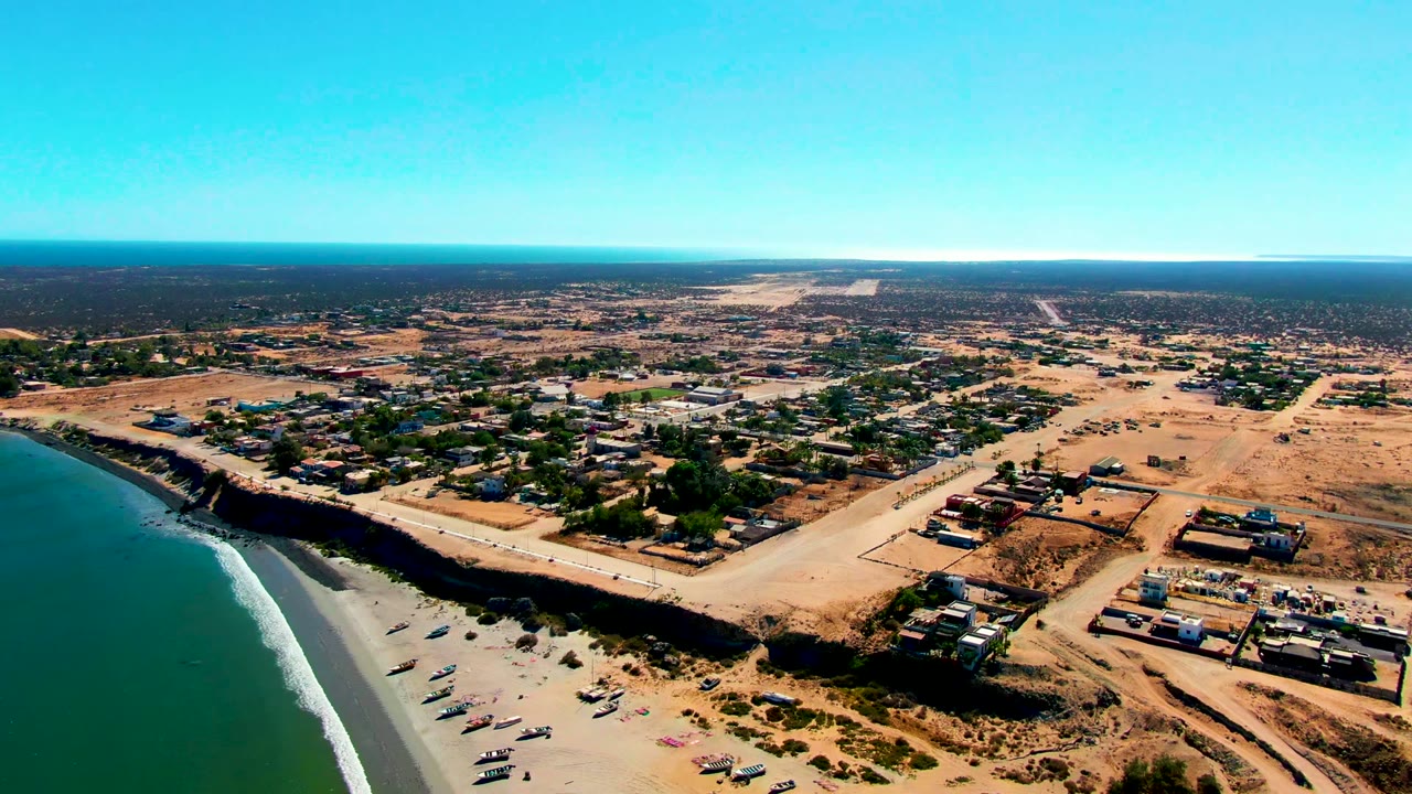 High aerial frame of San Juanico's ocean edge and beach, with a line of small boats near the shore, scattered structures beyond the road, dry terrain, and no visible people.