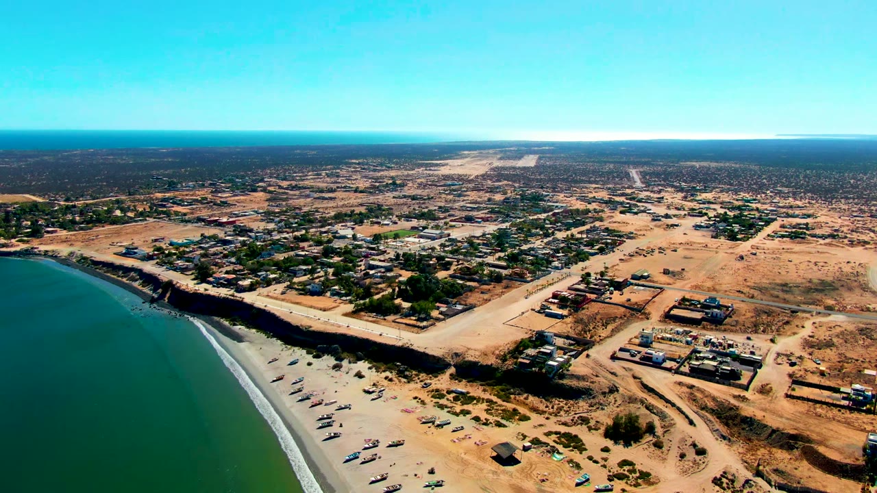 High aerial view over San Juanico, Baja California Sur, with green-blue ocean at left, a sandy beach below, dry lots and low buildings inland, and no visible people.