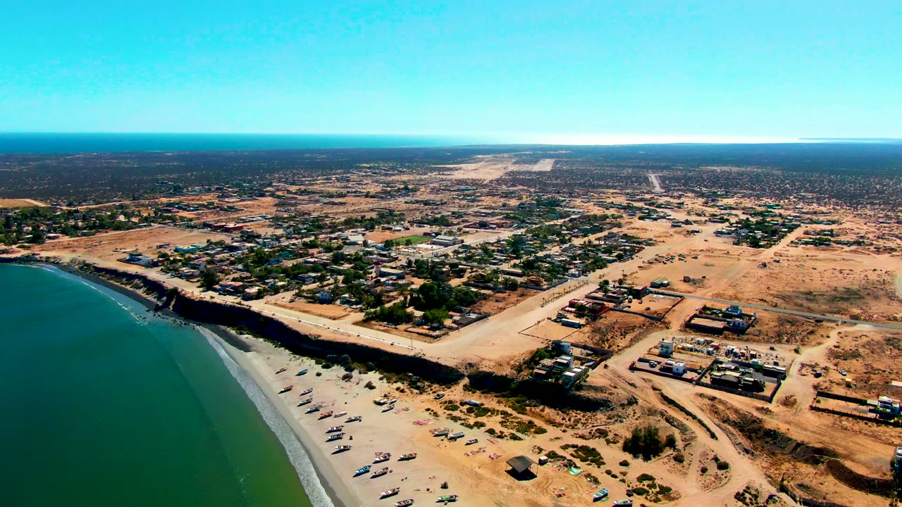 Drone view of a curved beach beside dry lots and low buildings.