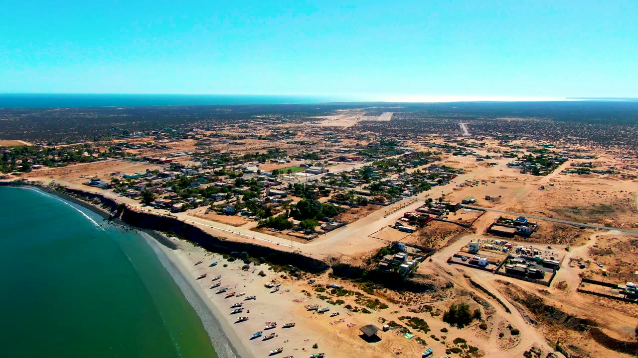 Wide drone view over San Juanico with the ocean occupying the left side, narrow surf on the beach, scattered buildings and vehicles inland, and no visible people.