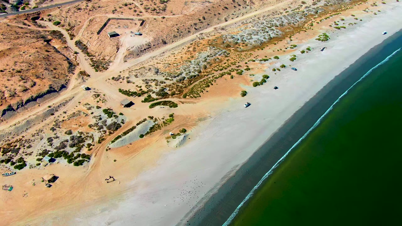 A high aerial frame over San Juanico, Baja California Sur shows a diagonal sandy shoreline, green ocean water, dirt roads, sparse shrubs, small structures, and no visible people.