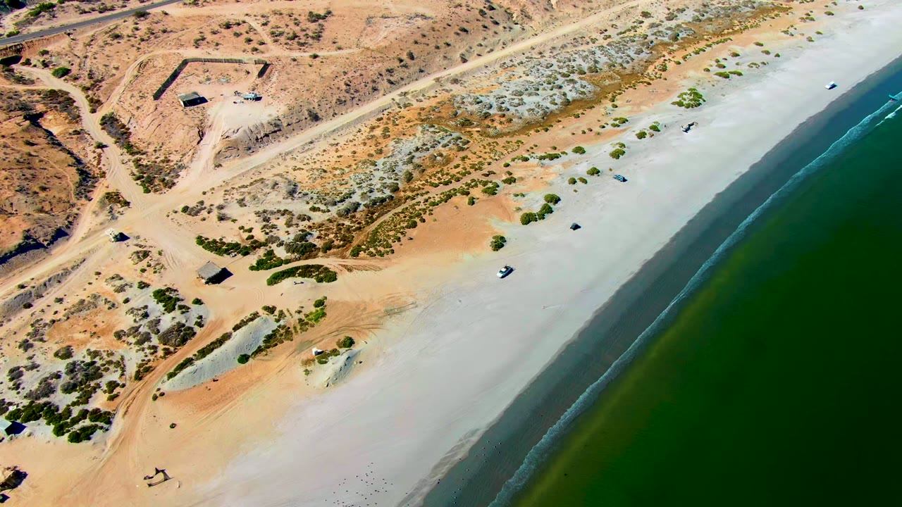 An aerial view of San Juanico shows the dirt road tracking above the beach, patchy shrubs on dunes, a narrow surf line, green water, and no visible people.