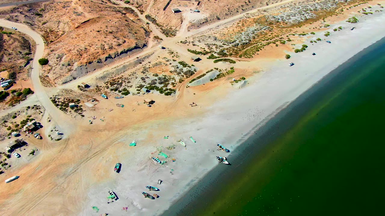 High aerial view over San Juanico, Baja California Sur, showing pale sand, green shoreline water, scattered small boats on the beach, dirt tracks, dry rocky terrain, and no visible people.