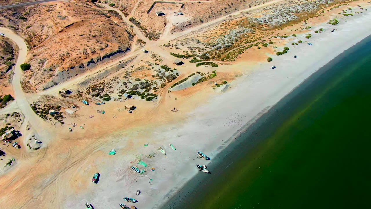Aerial coastline frame with boats on sand and dry hills beside green water.