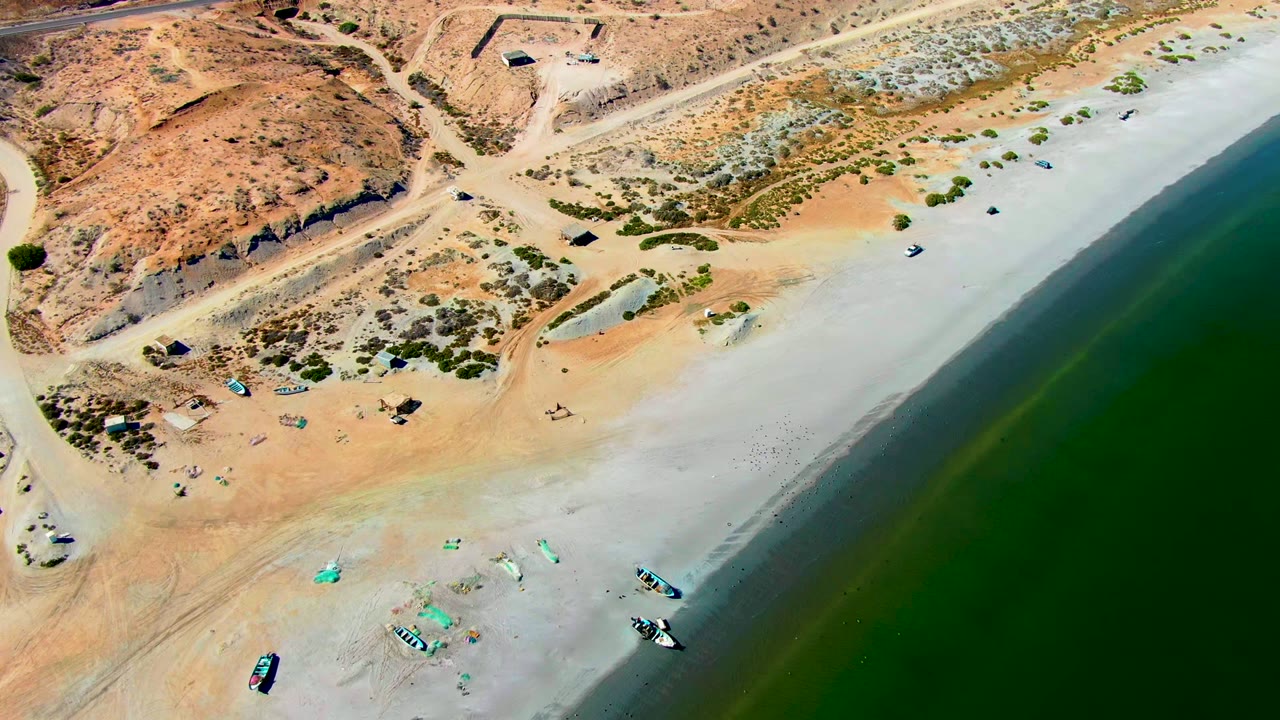 San Juanico aerial still showing a broad strip of pale beach, boats clustered near the lower sand, a small inland sandy depression, dirt roads, rocky desert ground, and no visible people.