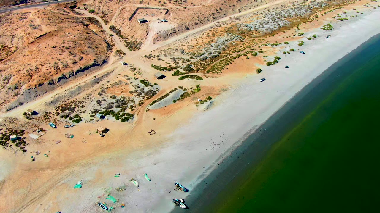 Aerial beach view with boats near the lower edge, green water to the right, and desert tracks above.