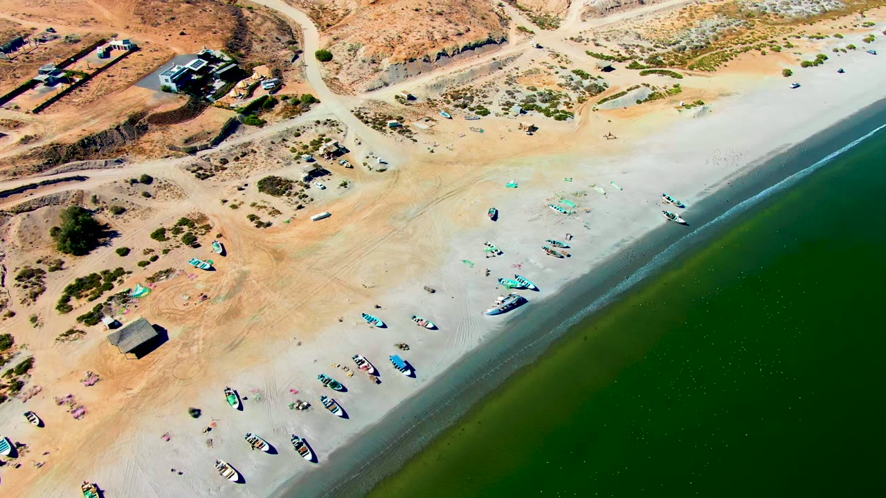Overhead view of San Juanico beach with boats on pale sand, green water along the right edge, a dirt road above the shore, and no visible people.