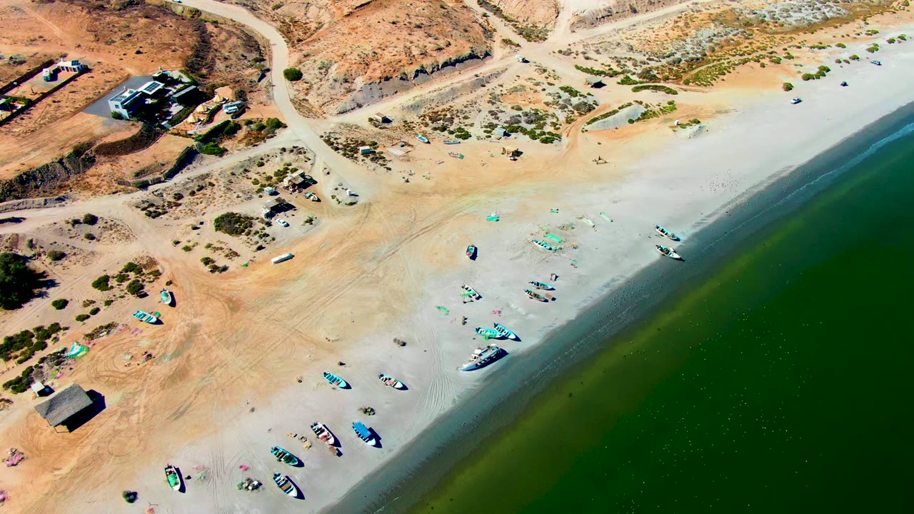High-angle view of San Juanico beach with the road crossing the upper frame, small boats near the lower sand, calm green water to the right, and no visible people.