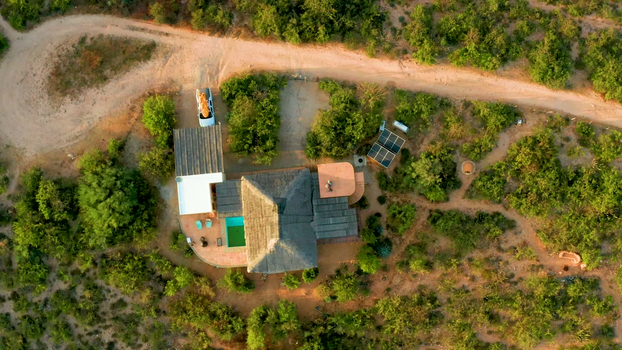 Overhead frame in Cabo Pulmo, Baja California Sur showing a rural house compound below a pale dirt road, surrounded by dense green brush, with no visible people.