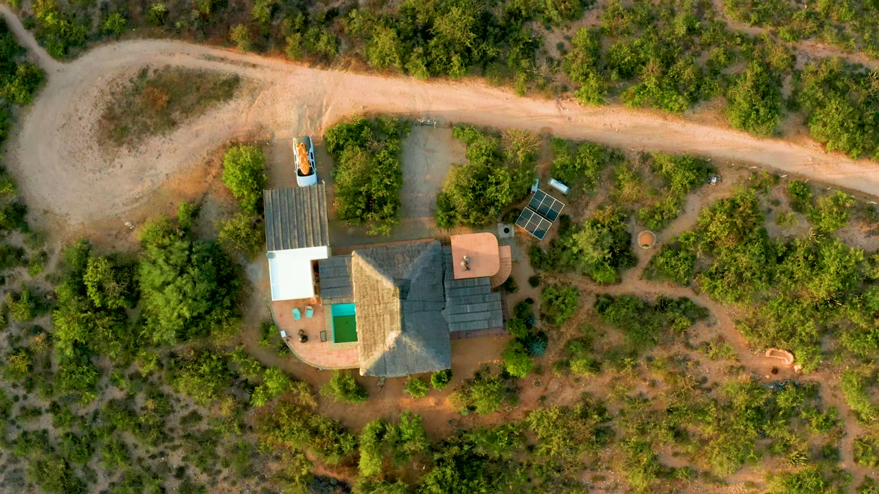 A high overhead frame in Cabo Pulmo shows the rural house at lower left, the dirt road at top, patches of brush across the land, and no visible people.