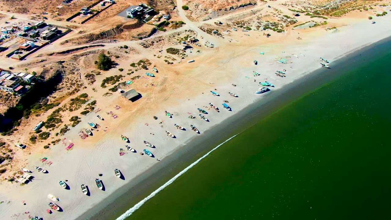 Aerial frame over San Juanico, Baja California Sur showing green water at lower right, a thin surf line, fishing boats on wet and dry sand, dune scrub, scattered structures, and no visible people.